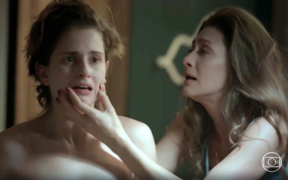A atriz Carol Duarte com cabelos cortados se olhando no espelho, e Maria Fernanda Cândida com mão no queixo dela, ambas com expressão de desespero