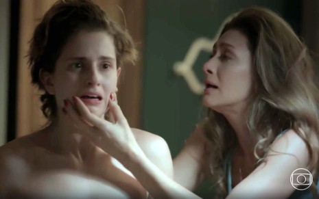 A atriz Carol Duarte com cabelos cortados se olhando no espelho, e Maria Fernanda Cândida com mão no queixo dela, ambas com expressão de desespero