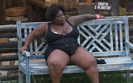 Jojo Todynho está sentada em um banco na área dos animais; a peoa usa maiô preto; ela está sentada com as pernas e braços abertos e olha para o lado
