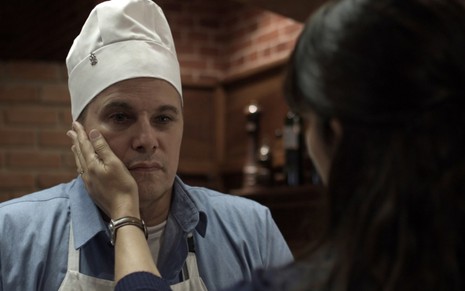 Edson Celulari com um chapéu de cozinheiro, enquanto uma mulher que está de costas para a câmera coloca a mão em seu rosto