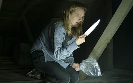 Agachada em um porão, Elisabeth Moss segura uma faca em uma mão e um saco plástico em outra em cena de O Homem Invisível (2020)
