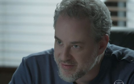 Dan Stulbach caracterizado como Eugênio em A Força do Querer: de camiseta preta, personagem está sentado e olha com surpresa para alguém fora do quadro