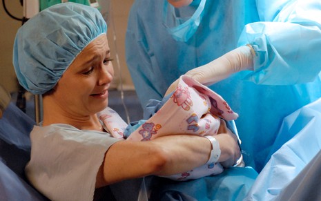 Deitada num leito de hospital, Julia Lemmertz segura um bebê recém-nascido caracterizada como a Esther em cena de Fina Estampa