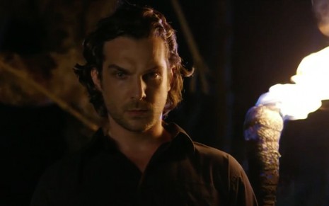 O ator Igor Rickli em uma tomada noturna, com uma tocha acesa ao fundo, caracterizado como Alberto em Flor do Caribe