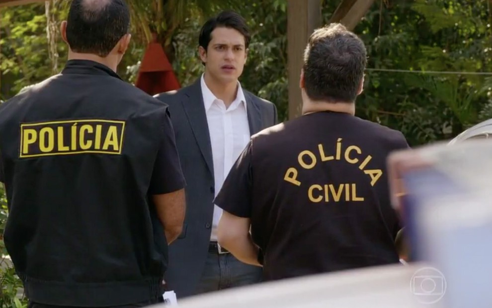 O ator Raphael Vianna com expressão de surpresa como Hélio com dois policias de costas, cujas jaquetas lê-se "polícia civil" em cena de Flor do Caribe