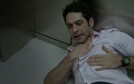 O ator João Baldasserini deitado no chão de um elevador, leva a mão à garganta e ao peito, como se tivesse falta de ar caracterizado como o Beto em cena de Haja Coração