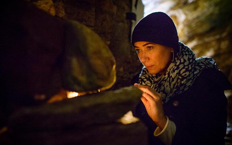 A atriz Lilia Cabral como Maria Marta está em uma caverna, com uma touca preta e um cachecol estampado, olhando para dentro de uma pedra em que brilha uma gema em cena de Império