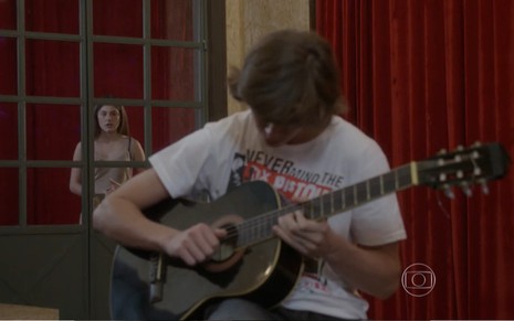 Rafael Vitti grava com camiseta estampada e violão na mão e Bruna Hamú o observa pela porta de vidro em cena de Malhação