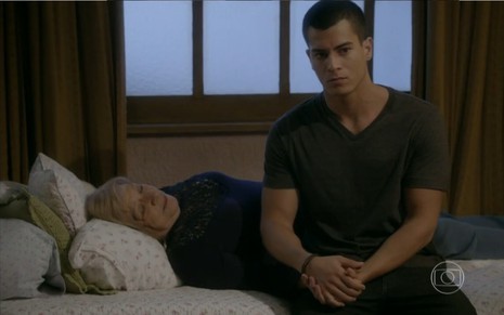 Iná de Carvalho grava de blusa azul escura deitada em uma cama com Arthur Aguiar sentado ao seu lado em cena de Malhação