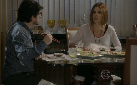 Guilherme Piva grava cena com Helena Fernandes, que usa camiseta de manga longa branca; eles comem em mesa um jantar japonês