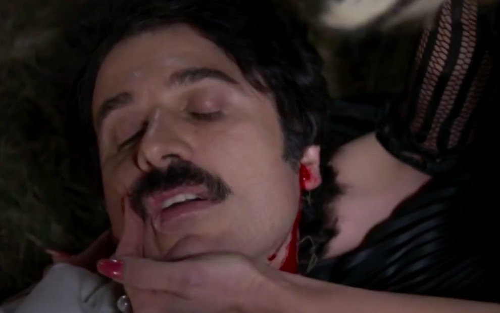 O ator Eriberto Leão tem o rosto segurado pela mão de Flávia Alessandra, há sangue descendo pelo seu pescoço, caracterizado como Ernesto em cena de Êta Mundo Bom!