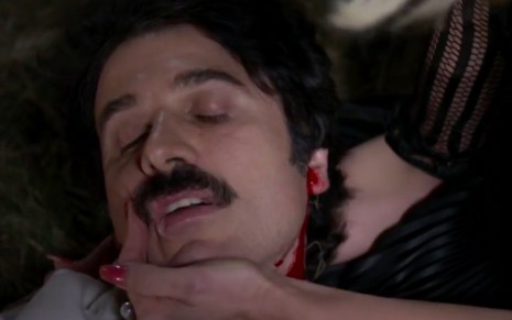 O ator Eriberto Leão tem o rosto segurado pela mão de Flávia Alessandra, há sangue descendo pelo seu pescoço, caracterizado como Ernesto em cena de Êta Mundo Bom!