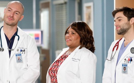 Na porta de um quarto de hospital estão, vestidos com aventais de médicos, Richard Flood, Chandra Wilson e Jake Borrelli, em Grey's Anatomy