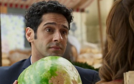 Beto (João Baldasserini) faz cara de pidão e segura uma melancia em frente a Tancinha (Mariana Ximenes) em Haja Coração