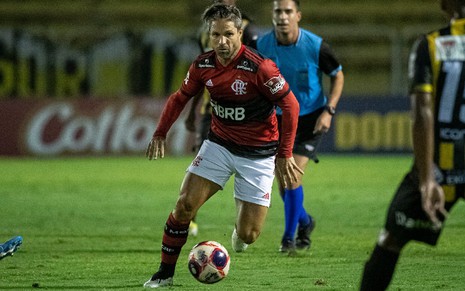 Diego com a camisa vermelha e preta e calção branco correndo com a bola sendo observado pelo juiz ao fundo