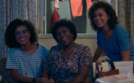 As atrizes Heslaine Vieira, Ju Colombo e Roberta Santiago sorriem em frente à televisão em cena de Malhação