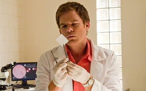 O ator Michael C. Hall, usando um avental branco, examina um pedaço de papel em cena de Dexter (2006-2013)