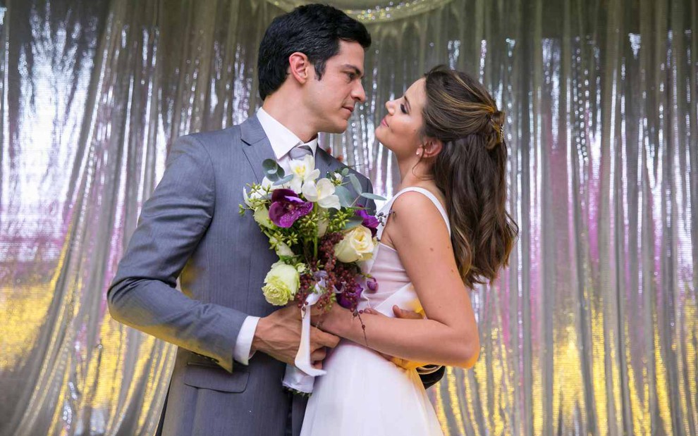 Mateus Solano veste um terno cinza e Camila Queiroz está de vestido branco com um buquê na mão. Eles estão casando-se.