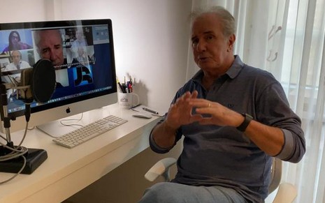 Com camisa social azul, Celso Freitas está sentado diante de um computador e de um microfone profissional