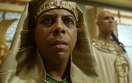 O ator André Ramiro está sentado em um trono dentro de um palácio egípcio com as roupas de um faraó na pele de Amenemhat 3º em cena de Gênesis