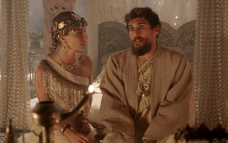 Maria Joana e Felipe Roque em cena de Gênesis: caracterizados como Enlila e Ibbi-Sim, atores estão sentados em uma cama e olham entre si
