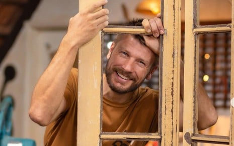 Rodrigo Hilbert sorrindo por trás de uma porta com camisa mostarda, em foto de divulgação do Tempero de Família