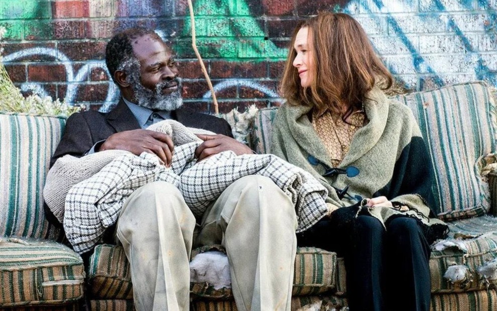Sentados em um sofá rasgado, Djimon Hounsou e Renée Zellweger se olham sorrindo em cena de Somos Todos Iguais (2017)