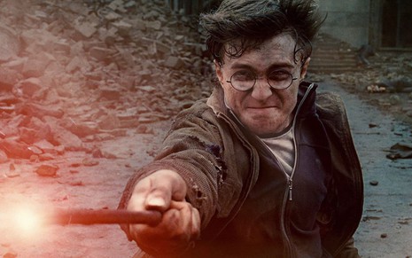 Caído no chão e com expressão de esforço, Daniel Radcliffe aponta varinha e dispara um feitiço vermelho