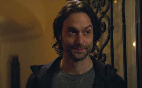 Na porta de uma mansão, o ator Chris D'Elia aparece de barba, camisa cinza e blusa escura em cena da série You