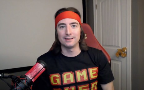 Keith Grill em vídeo publicado no YouTube: sentado, youtuber usa faixa vermelha, camiseta preta e está do lado de um microfone de mesa vermelho
