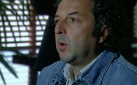 O ator Luiz Ramalho como Baiano em A Favorita; ele está sentado, de lado, com a boca semiaberta ernquanto conversa durante um depoimento da cena