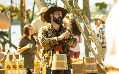 O ator Juliano Cazarré como Alcides em Pantanal; ele está de chapéu batendo palmas em volta de um barril de cachaça