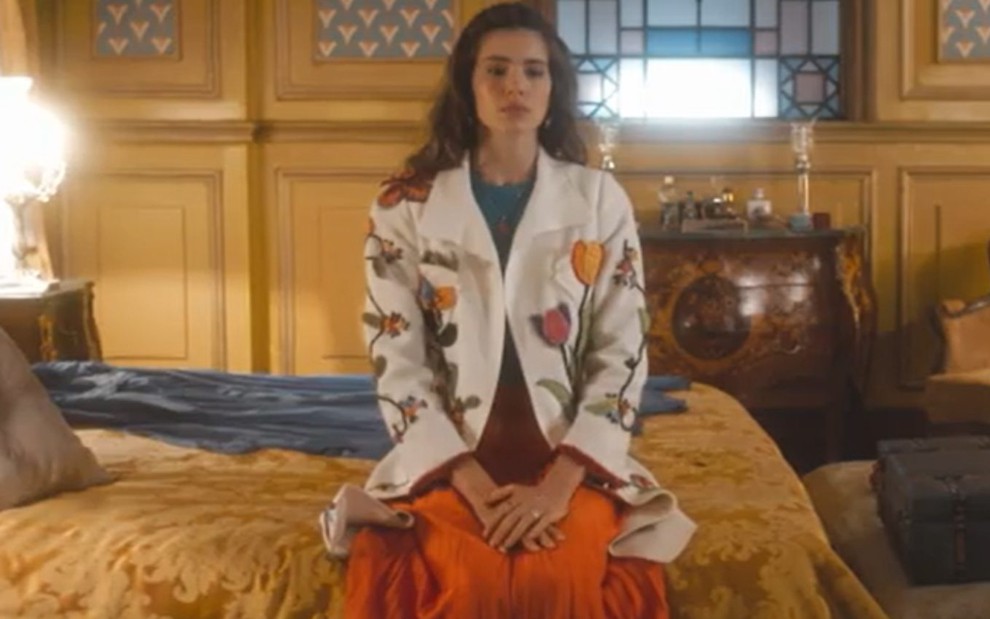 A atriz Camila Queiroz sentada numa cama, com roupa de festa e expressão triste em cena de Amor Perfeito