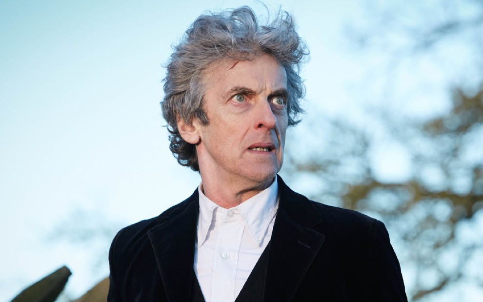 Doctor (Peter Capaldi) de camisa branca e jaqueta preta olhando para a direita com cenário de floresta desfocada ao fundo em cena de Doctor Who