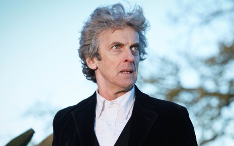 Doctor (Peter Capaldi) de camisa branca e jaqueta preta olhando para a direita com cenário de floresta desfocada ao fundo em cena de Doctor Who
