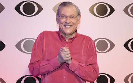 O apresentador Milton Neves usa blusa avermelhada e posa em frente a um painel com logotipo da Band