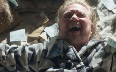 O ator Kiko Mascarenhas como Duarte/Bob Wright em Cara e Coragem; ele está deitado, rodeado de notas de dinheiro, com um sorriso no rosto
