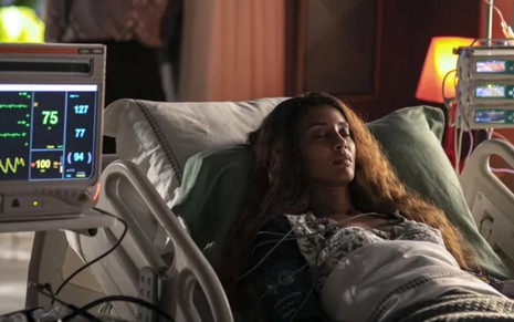A atriz Taís Araujo deitada em maca de hospital, monitorada por aparelhos, em cena de Cara e Coragem