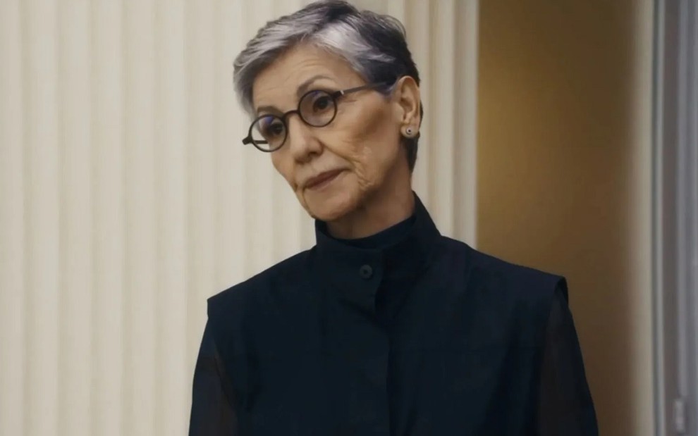 Cassia Kis com expressão séria, óculos e blusa preta de gola alta em cena como Cidália na novela Travessia