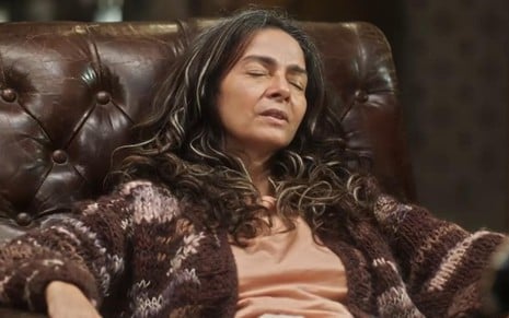 A atriz Claudia Ohana de olhos fechados, sentada em poltrona com expressão cansada, em cena de Vai na Fé