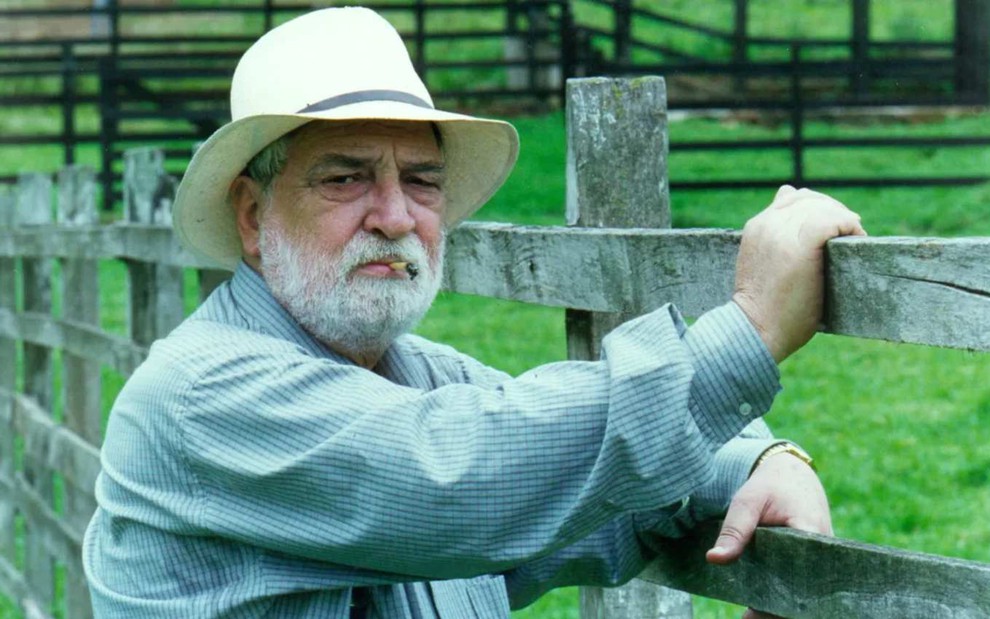 O ator Lafayette Galvão (1931-2019) caracterizado como seu personagem em Corpo Dourado, com camisa azul, chapéu branco, cigarro na boca, com as mãos em uma cerca, num pasto