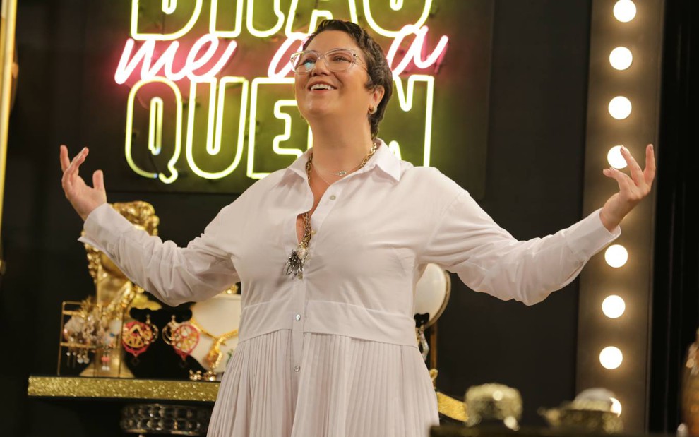 A cantora Maria Rita de vestido branco, sorrindo, em estúdio do programa Drag Me as a Queen, do canal E!