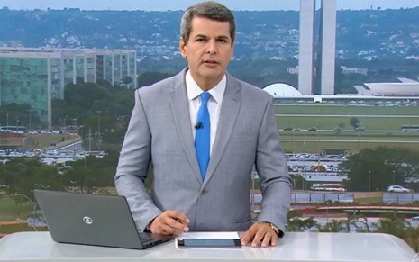 O jornalista Fábio William com expressão séria, de terno e gravata, na bancada do DF1, com paisagem da cidade de Brasília no plano de fundo
