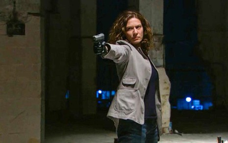A atriz Patricia Pillar como Flora em A Favorita; ela está olhando para o lado e apontando uma arma com cara de crueldade