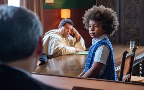 Levi Asaf caracterizado como Marcelino; o ator usa uma camisa branca e um colete xadrez azul. Ele está no meio de um tribunal, cercado por Babu Santana e Chico Pelúcio, e parece tenso.