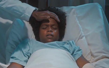 O ator Levi Asaf está deitado em um leito com alguém colocado a mão na sua testa para medir temperatura em Amor Perfeito