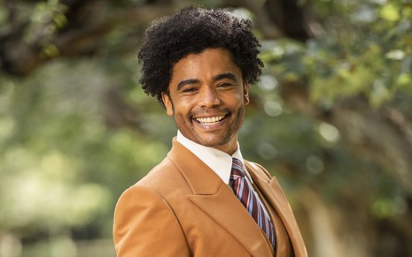 Diogo Almeida posa com terno e gravata em um jardim como o médico Orlando da novela Amor Perfeito, da Globo