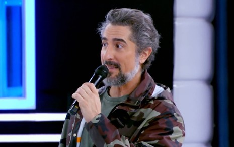 Marcos Mion usa uma blusa verde e uma jaqueta marrom; apresentador segura o microfone na direção da boca