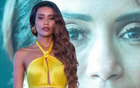 A atriz Taís Araujo está com vestido amarelo de festa em frente a um telão no qual aparece foto de seu rosto em cena de Cara e Coragem