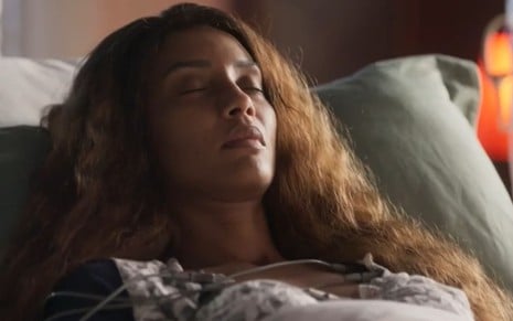 Taís Araujo caracterizada como Clarice; cabelos lisos e grossos estão soltos, ao redor da cama, em cena de Cara e Coragem